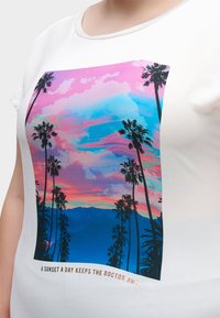 Wit overhemd met een kleurrijke print van zonsondergang en palmbomen, met de tekst "Een zonsondergang per dag houdt de dokter weg" onder de afbeelding.