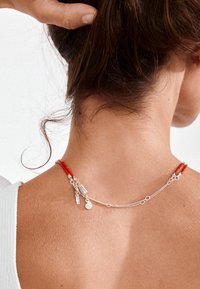 Cuello de mujer con cabello castaño recogido, lleva un delicado collar de plata con cordón rojo y pequeños colgantes.