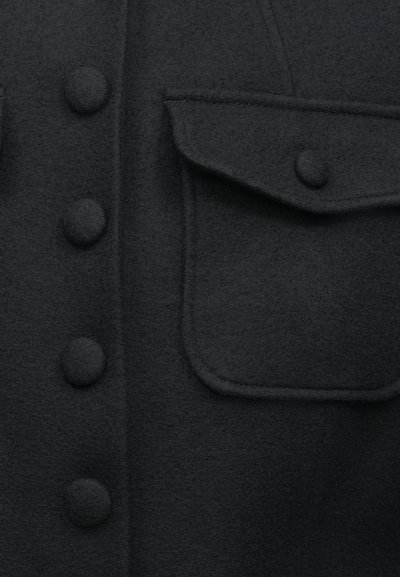 Sort fleece-skjorte med store runde knapper og en brystlomme med klap. Teksturen virker blød og let dunet.