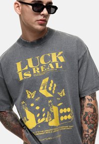 Szara koszulka z krótkim rękawem, z żółtym graficznym tekstem oraz ilustracjami kostek i motyli, o luźnym kroju i okrągłym dekolcie.