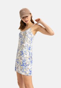 Vestido de estampado floral en blanco con flores azules y verdes, con tirantes finos y un corte holgado. Combinado con una gorra beige.
