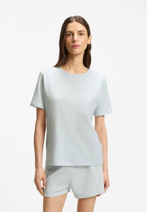 Kvinde stående vendt fremad iført en ensfarvet lyseblå t-shirt med korte ærmer og matchende shorts mod en hvid baggrund.
