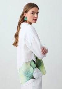 Camicia in lino bianco con grande stampa di foglie verdi e nere sul lato, colletto classico e vestibilità rilassata. Modella che indossa orecchini turchesi.