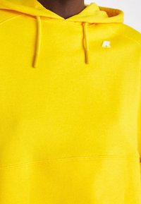 Personne portant un sweat à capuche jaune vif avec des cordons et un petit logo blanc sur la partie supérieure de la poitrine près de l'épaule gauche.