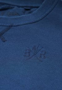 Blauer gestrickter Pullover mit einem gerippten Kragen und aufgestickten gekreuzten Buchstaben "B" und "R" im Brustbereich.