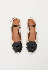 Beige slingback flats with a black leather flower accent on the toe. Smooth texture with an open design and brown interior.