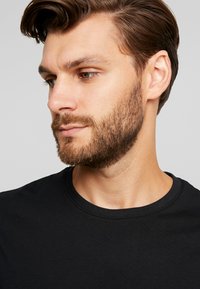 Black round-neck t-shirt on a man with light skin, short brown hair, and a beard. The fabric appears smooth and fitted around the neck.