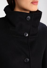 Svart ullrock med hög krage, med tre stora svarta knappar och en slät textur. Närbild som fokuserar på halsringningen.