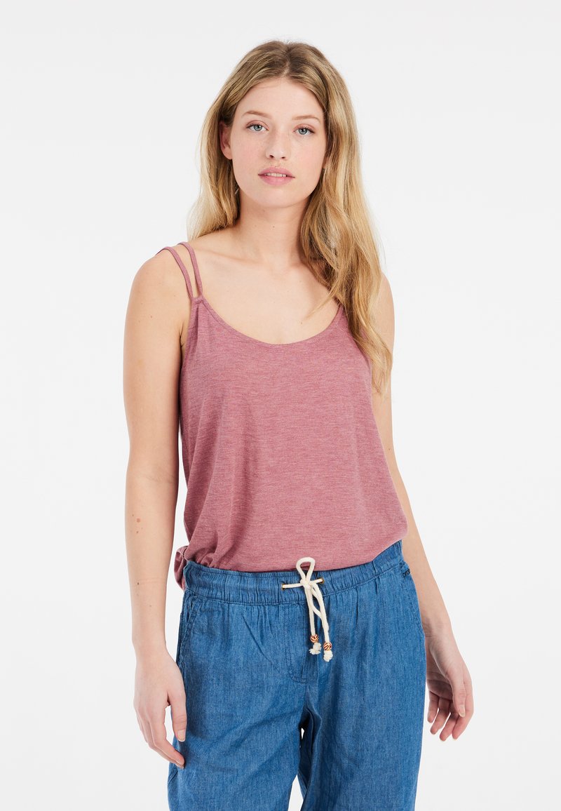 Joven mujer con el pelo largo y rubio, que lleva una camiseta rosa sin mangas y pantalones azules con cordón, de pie frente a un fondo blanco.