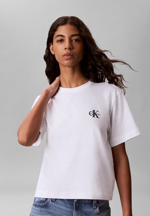 Ung kvinde med langt krøllet hår iført hvid Calvin Klein t-shirt og blå jeans, står mod grå baggrund, med hånden rørende ved halsen.