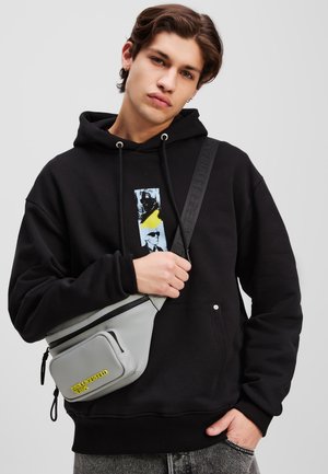 Schwarzer Hoodie mit Grafikdruck, graue Umhängetasche mit gelbem Logo und graue Jeans. Die Tasche verfügt über Reißverschlüsse und eine strukturierte Oberfläche.