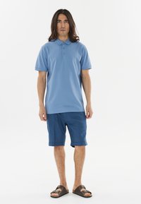 Vaaleansininen polo-paita, jossa on kaulus ja kolme nappia, yhdistettynä sinisiin shortseihin. Sandalet, joissa on ruskea hihna ja tumma pohja.