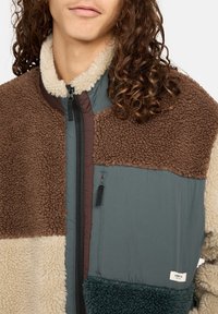 Veste en polaire texturée avec des panneaux marron, beige et vert ; fermeture éclair et col doux ; tissu durable avec des motifs contrastés.