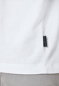 T-shirt bianco in cotone con una texture liscia. Presenta un piccolo etichetta nera "AllSaints" sul lato. L'orlo è cucito e uniforme.