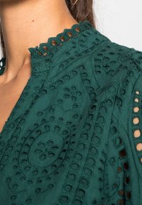 Mörkgrön blus med intrikata broderier, vågig halsringning och utskärningar. Tyget verkar ha en texturerad yta med ett mönstrat design.