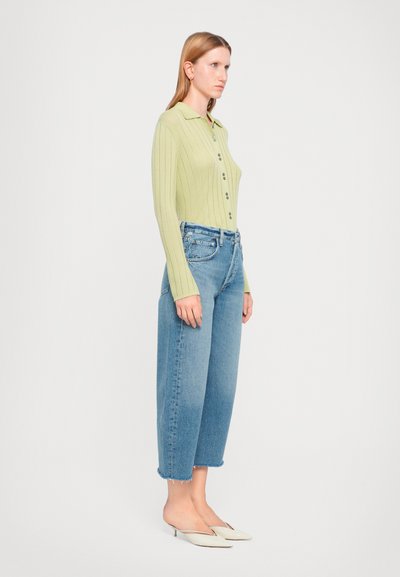 Pull léger vert clair à côtes à demi-boutonnage, associé à un jean en denim bleu surdimensionné avec des ourlets effilochés et des chaussures blanches à enfiler.