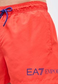 Κόκκινα μαγιό βερμούδα με μπλε κορδόνι και τυπωμένο το λογότυπο EA7 Emporio Armani κοντά στο κάτω τελείωμα στο αριστερό πόδι.