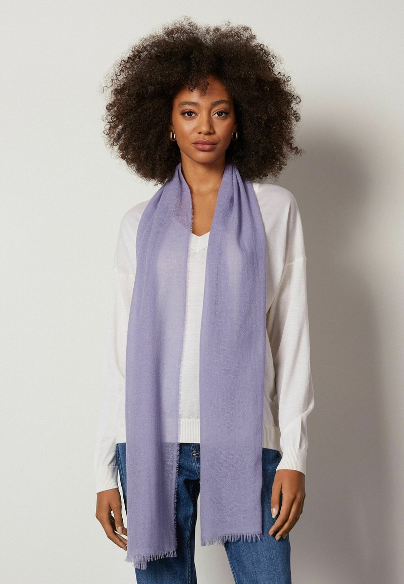 Lyse lilla tørklæde lavet af blødt materiale med frynset kanter, der hænger elegant om halsen, båret over en hvid top og jeans.
