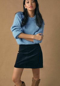 Pull côtelé bleu clair à manches retroussées, associé à une mini-jupe en velours côtelé noire. Des bottes hauteur genou couleur beige complètent la tenue.