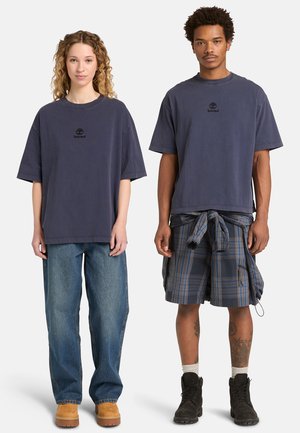 Zwei Personen tragen marineblaue Timberland-T-Shirts; eine kombiniert es mit weiten Jeans und beigen Stiefeln, die andere mit karierter Shorts, Stiefeln und einem gebundenen Hemd.