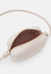 Světlá béžová kožená křížová taška se zlatým zipem, zaobleného tvaru a hladkou texturou. Uvnitř má hnědou podšívku a detail loga.