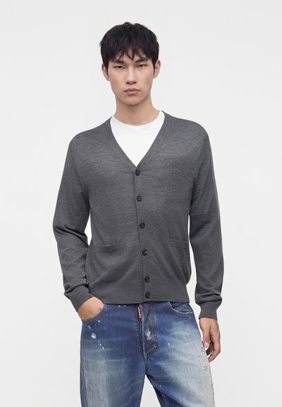 Jeune homme portant un cardigan boutonné gris foncé sur une chemise blanche et un jean bleu déchiré, la main gauche dans la poche.