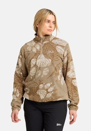Vrouw met een beige en bruin gebreide fleece met rits bij de hals en zwarte broek met logo, staand tegen een eenvoudige lichte achtergrond.
