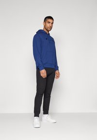 Sweat à capuche bleu avec poche avant, pantalon noir slim-fit et baskets blanches. Design simple avec des poignets côtelés et un détail de logo.