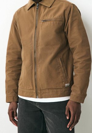 Homme portant une veste marron zippée avec des poches zippées sur la poitrine et les côtés, chemise blanche en dessous et jean foncé sur fond uni.