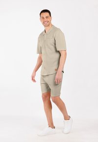 Lachende man die loopt, gekleed in een beige overhemd met korte mouwen, bijpassende shorts en witte sneakers, tegen een effen witte achtergrond.