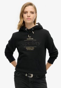 Sudadera con capucha negra, bolsillo frontal y cordón, con un logotipo en relieve en dorado. Combinada con unos jeans negros y un cinturón con hebilla plateada.