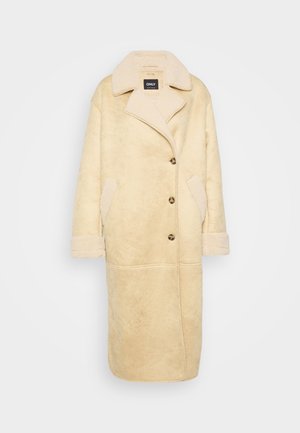 Beige nepbont jas met een schouderkraag, voorzien van grote knopen, zijzakken en textuurstof. Lange mouwen met omgeslagen manchetten.