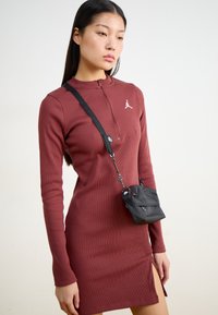 Abito lungo a maniche lunghe di colore marrone bordeaux con colletto a zip quarto di giro e uno spacco laterale. Una piccola borsa a tracolla nera è indossata sulla spalla.
