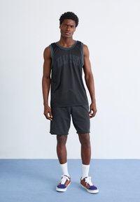 Fekete ujjatlan tank top kék díszítéssel és "ELLESSE" logóval, összeillő fekete rövidnadrággal és színes mintás sportcipővel párosítva. Fehér zoknik láthatóak.