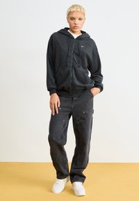 Felpa nera con zip e tasche frontali, indossata sopra una maglietta bianca, abbinata a pantaloni cargo neri larghi con zip e una vestibilità rilassata.