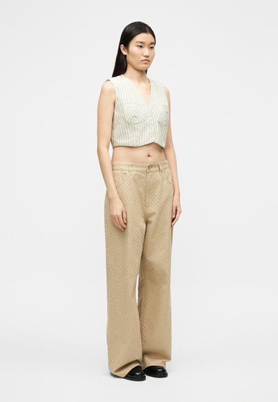 Γυναίκα που στέκεται φορώντας ένα ριγέ cropped γιλέκο χωρίς μανίκια και μπεζ παντελόνι με φαρδιά πόδια και μικρές οπές, συνδυασμένο με μαύρα παπούτσια.