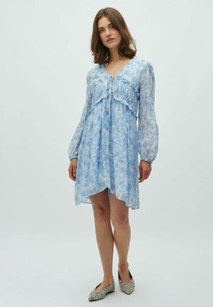 Vestido azul claro con estampado floral, escote en v, detalle fruncido en el busto, mangas largas abullonadas y dobladillo asimétrico. Combinado con zapatos planos estampados.