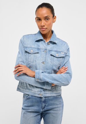 Frau mit verschränkten Armen, die eine hellblaue Jeansjacke und passende Jeans trägt, steht vor einem schlichten hellen Hintergrund.