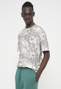 Γκρι μπλουζάκι με κοντά μανίκια και στρογγυλή λαιμόκοψη, με ποικιλόμορφο κολάζ εκτύπωση, σε συνδυασμό με πράσινη φόρμα με κίτρινο λογότυπο.