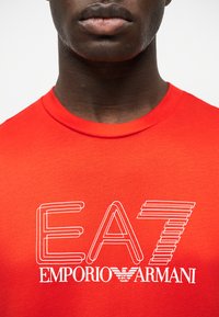 Άνδρας φοράει κόκκινο μπλουζάκι με λευκό λογότυπο "EA7 Emporio Armani" στο κέντρο του στήθους, μπροστά σε απλό λευκό φόντο.