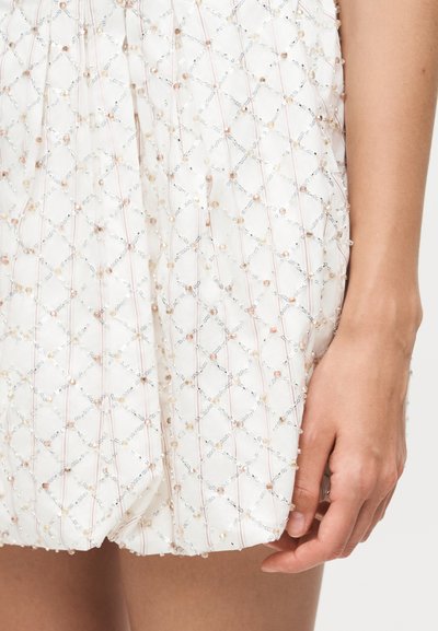 Λευκό ύφασμα διακοσμημένο με δίχτυ σε μοτίβο διαμαντιού από μικρές παγιέτες και χάντρες, εμφανίζεται κοντά στο χέρι και τον μηρό ενός ατόμου.