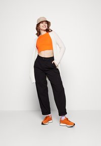 Narancssárga crop top, fehér nyitott kardigán, fekete cargo nadrág és narancssárga sportcipő rózsaszín zoknival. A bézs vödörsapka teszi teljessé a megjelenést.