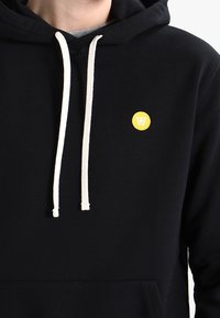 Svart huvtröja i mjukt tyg, med vita dragsko och en liten gul cirkulär logotyp på bröstet. Enkelt design, inga mönster.