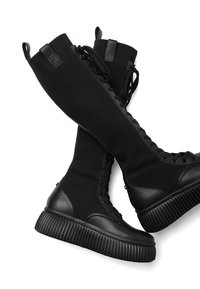 Bottes hautes noires à lacets avec tige en tissu côtelé, semelle épaisse à rainures, embout en cuir et languettes de traction sur fond blanc.