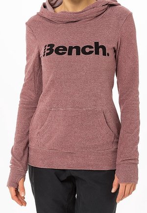 Femme portant un sweat à capuche mauve à manches longues avec poche avant et logo noir "Bench.", associé à un pantalon noir.