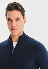 Granatowy sweter na zamek z wysokim kołnierzem, wykonany z gładkiej dzianiny. Posiada mały zamek i czyste linie. Jednokolorowy, z minimalnymi zdobieniami.