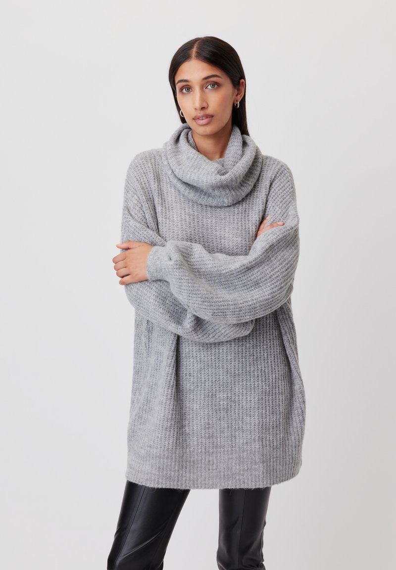 Jasnoszara, oversize sweter z golfem, teksturowana dzianina, długie rękawy i luźny krój, zestawiony z czarnymi skórzanymi spodniami.