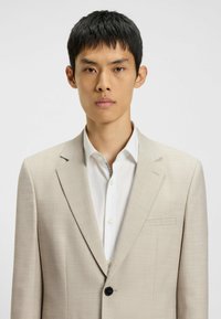 Beige blazer lavet af tekstureret stof, enkeltknaplukning, spidse revers og en lomme; båret over en hvid skjorte med krave.