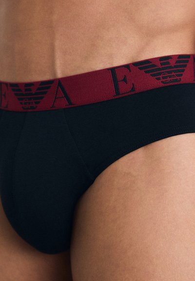 Schwarze Baumwollslips mit einem kastanienbraunen elastischen Bund, der Logo-Text und ein Adleremblem zeigt. Glatte Textur, figurbetontes Design.
