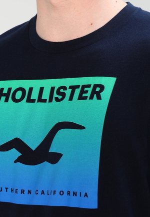 Zbliżenie granatowego T-shirtu z prostokątem o zielono-niebieskim gradiencie, czarną sylwetką ptaka i napisem "Hollister Southern California".